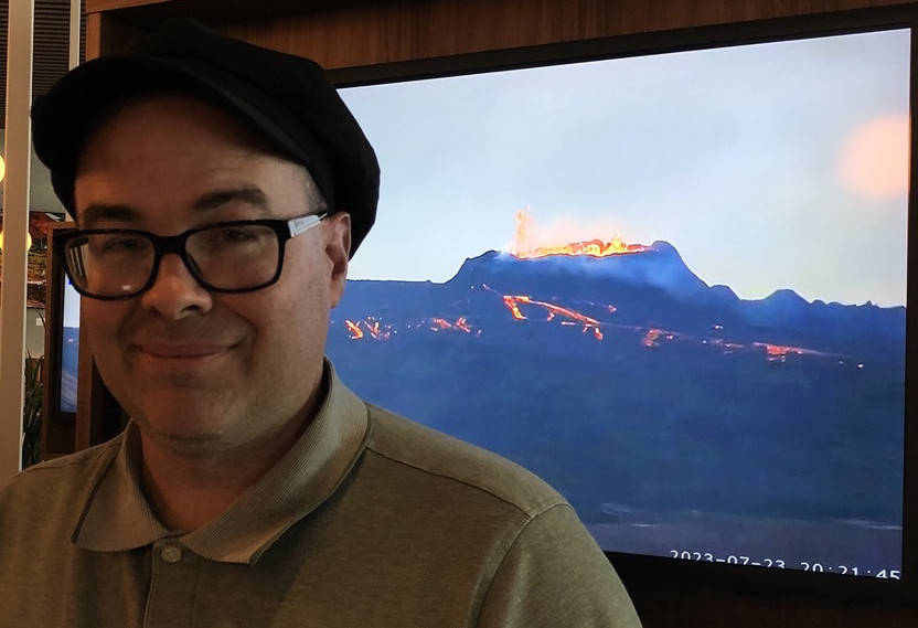 Toby sitting in front of a television which depicts a volcanic crater spewing lava, with lava flows in front of it.